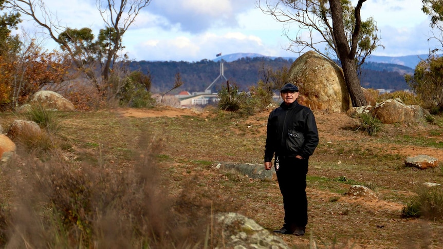 A man wearing stands in a clearing surrounded by rocky outcrops, with Parliament House visible in the distance.
