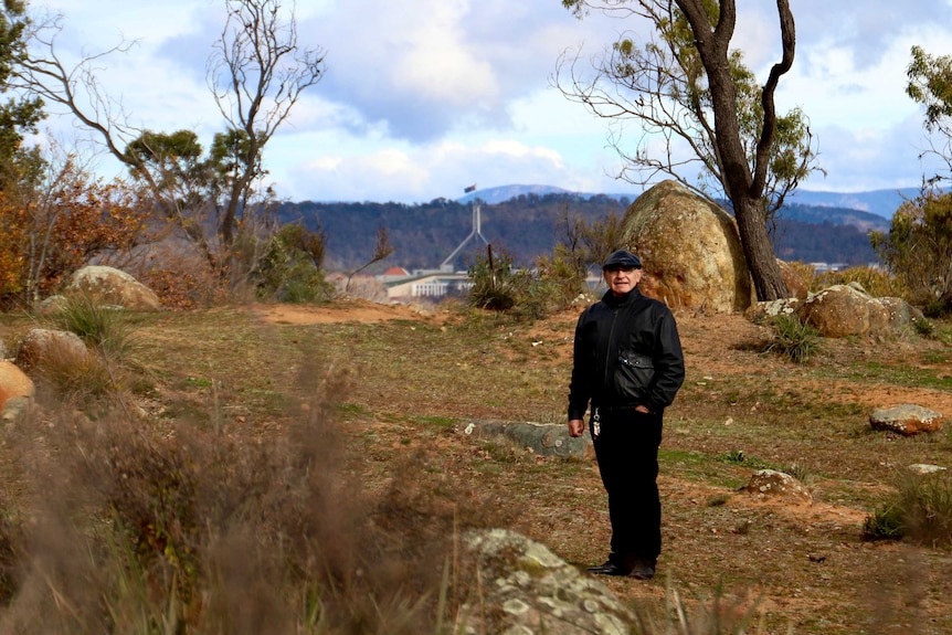 A man wearing stands in a clearing surrounded by rocky outcrops, with Parliament House visible in the distance.