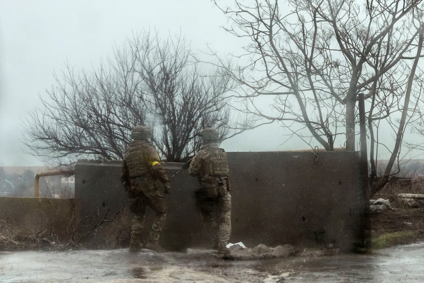 Two soldiers dressed in fatigues hold guns as they stand near a wall in the snow