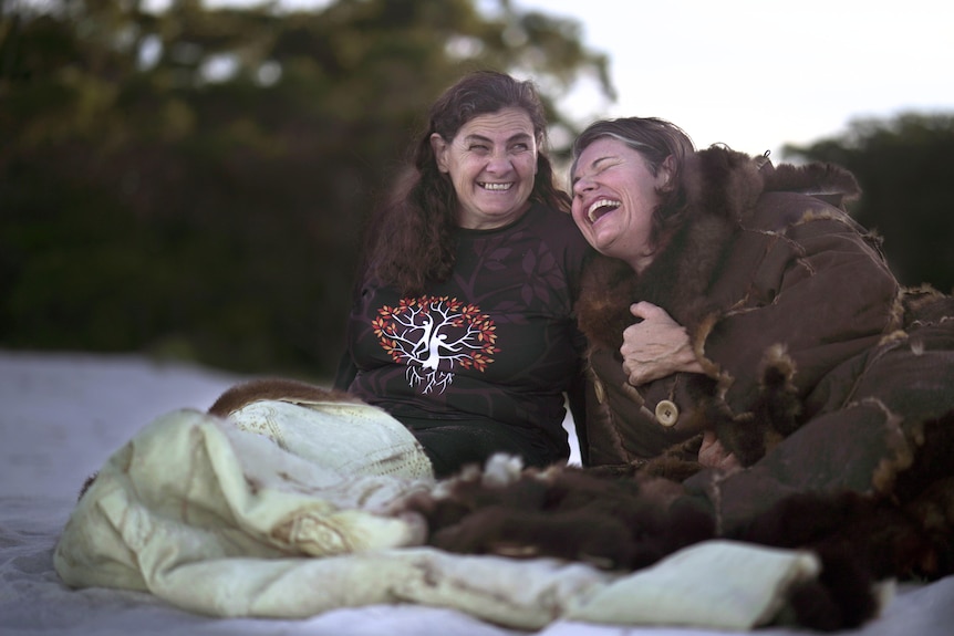 Two women laugh while sitting by a fire on the beach. One woman is wearing a large possum skin coat