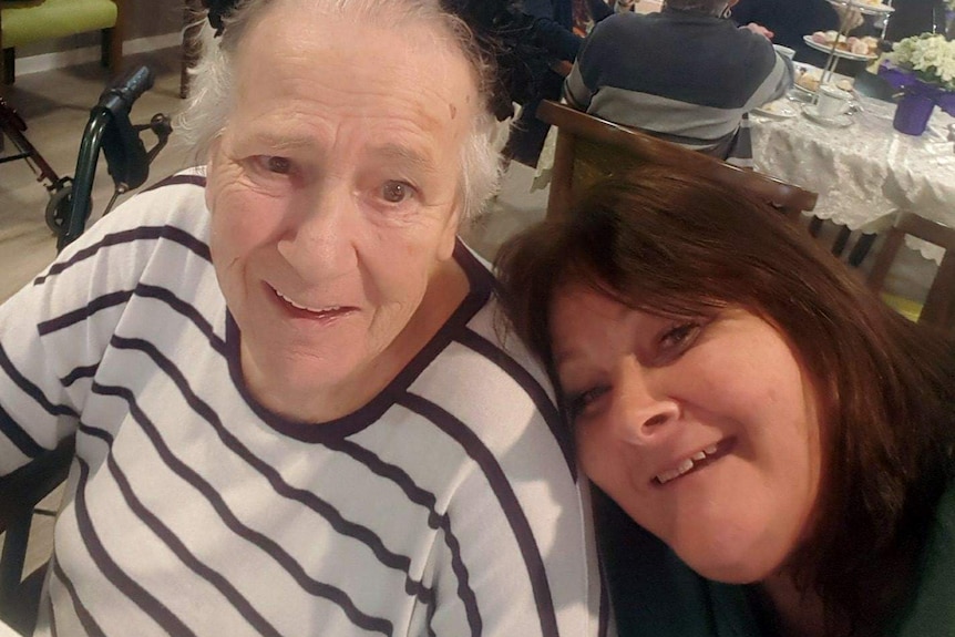 Older woman with striped shirt smiles with her daughter leaning on her shoulder smiling inside an aged care home dining room