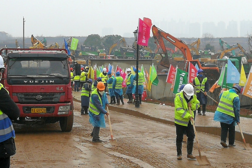 workers in florescent vests and helmets work near the entrance of a construction site in Wuhan with cranes in the background