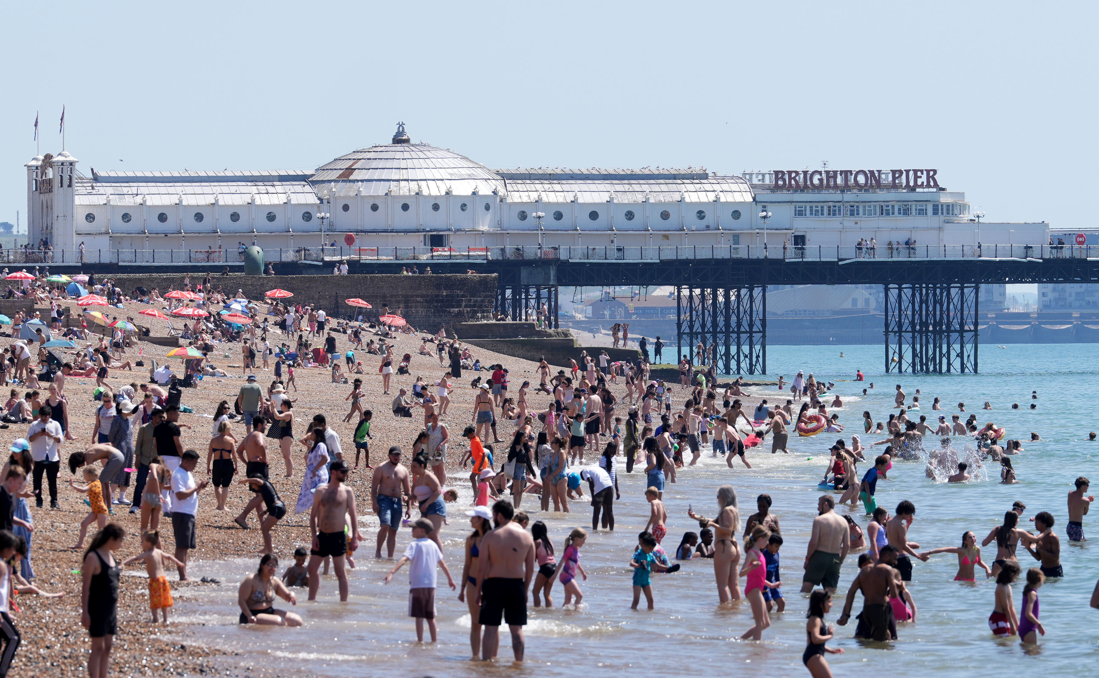 Hundreds of people dotted across a wide beach with a pier in the background, on a sunny day.