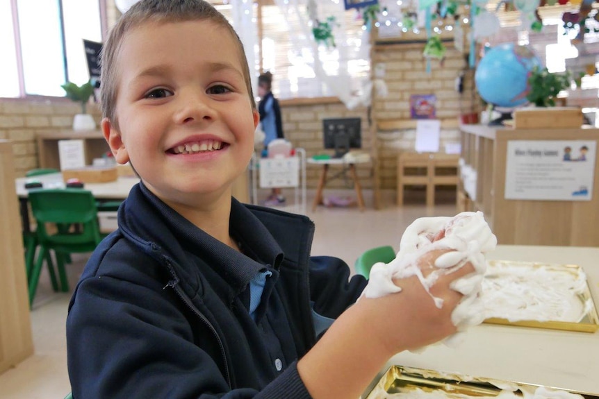 A boy smiles sitting at a table in a classroom grasping a white foam-type substance in his hands.