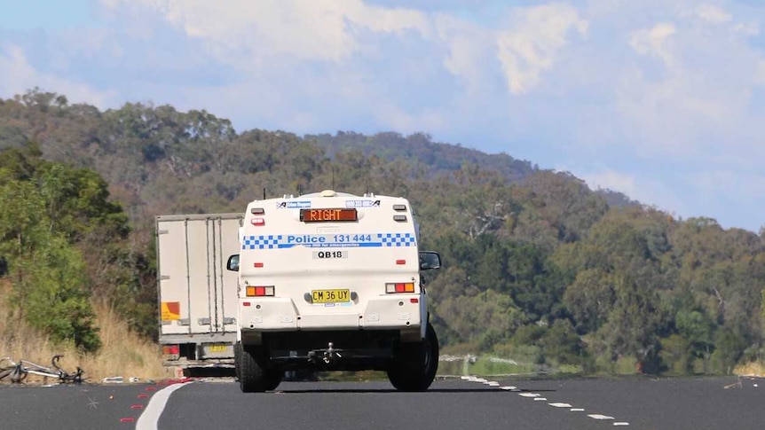 A bike lays on the side of the road with a white body bag beside it, with policeman and police van nearby.