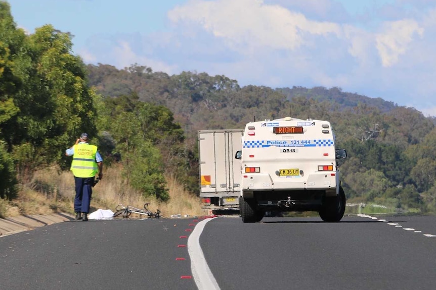 A bike lays on the side of the road with a white body bag beside it, with policeman and police van nearby.