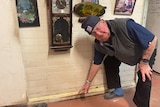 An old man bends down pointing to a crack between the wall and the floor of his home 