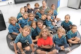 A group of pre primary students sit on the mat with their teacher smiling at the camera