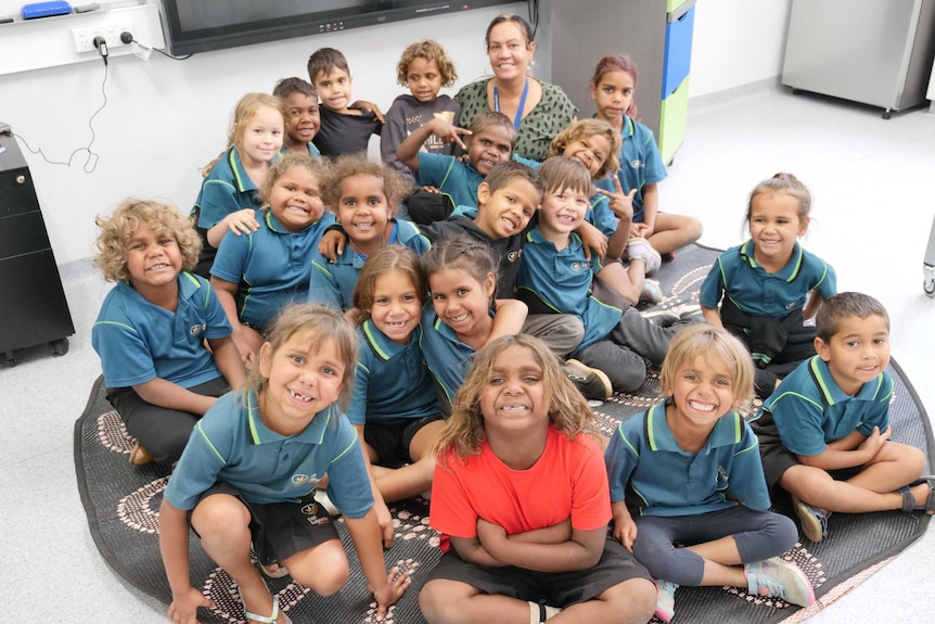 A group of pre primary students sit on the mat with their teacher smiling at the camera