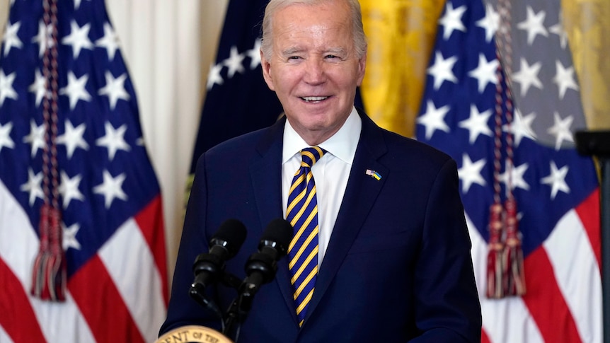 Joe Biden smiles while standing at a podium in front of US flags.