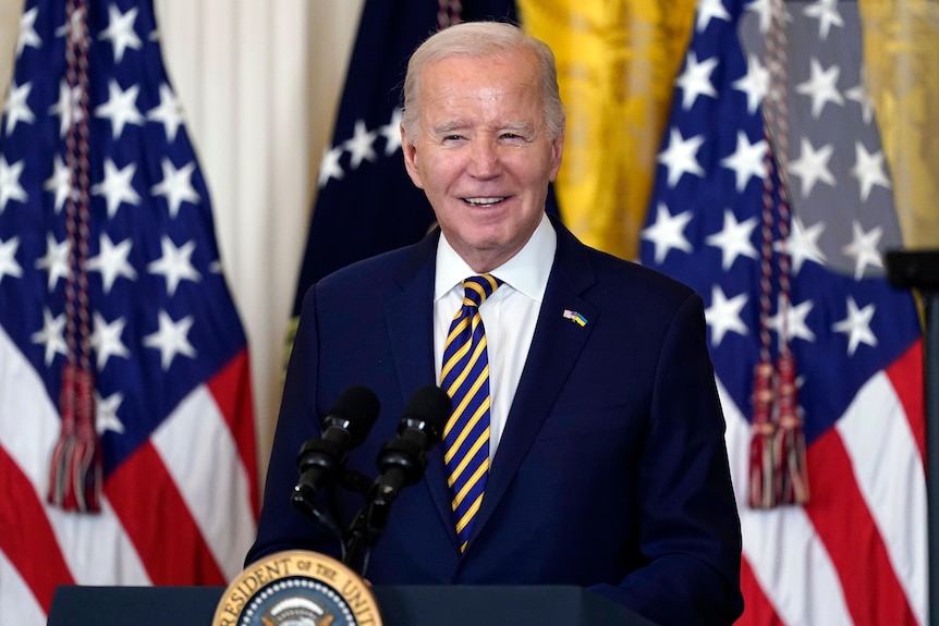 Joe Biden smiles while standing at a podium in front of US flags.