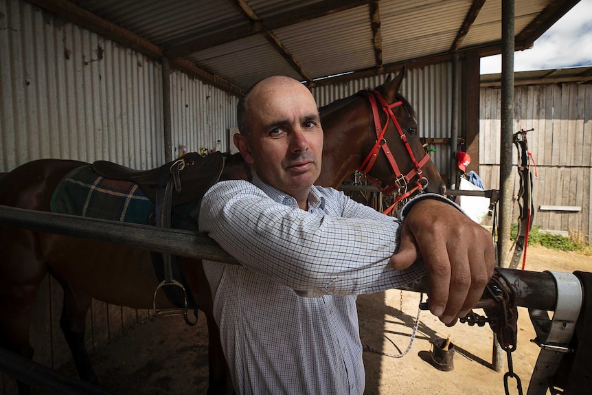 Horse trainer standing in a shed with a saddled horse behind him