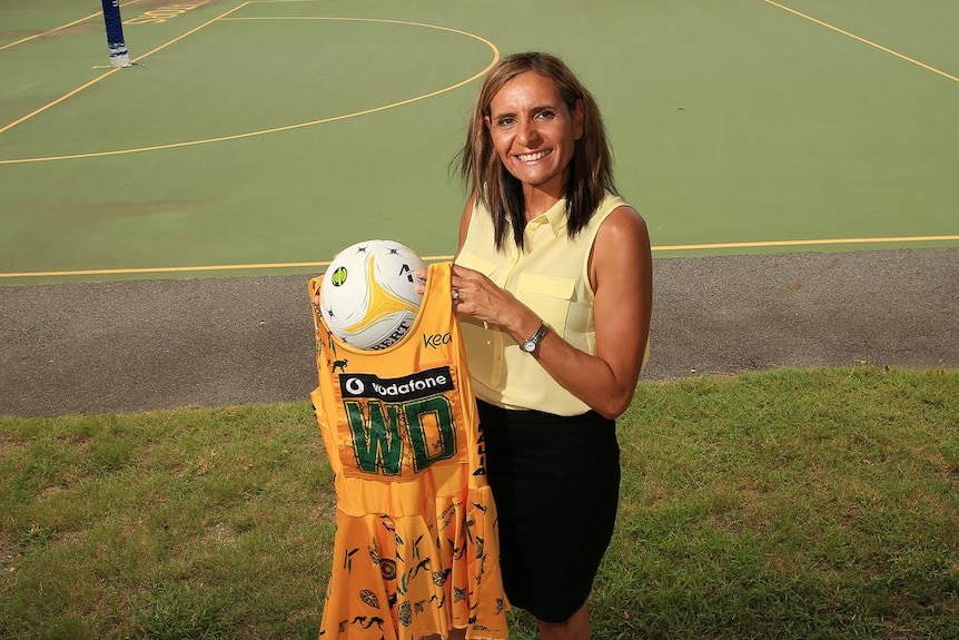 Sharon Finnan-White stands on the grass in front of a netball court, smiling as she holds her old Diamonds dress and a netball