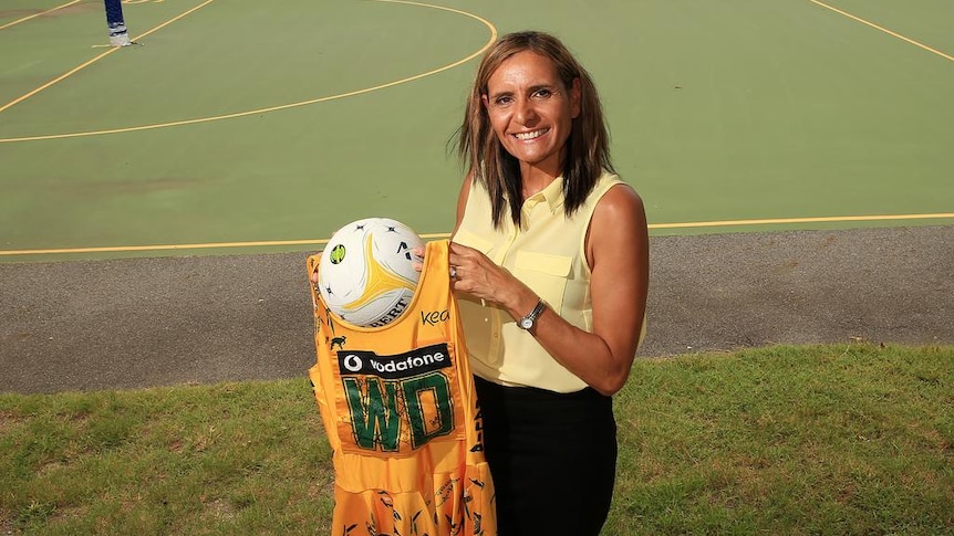 Sharon Finnan-White stands on the grass in front of a netball court, smiling as she holds her old Diamonds dress and a netball