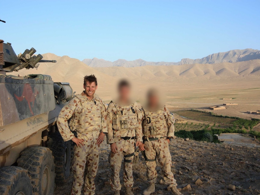 Three soldiers standing next to a tank in a valley  in front of large brown mountains