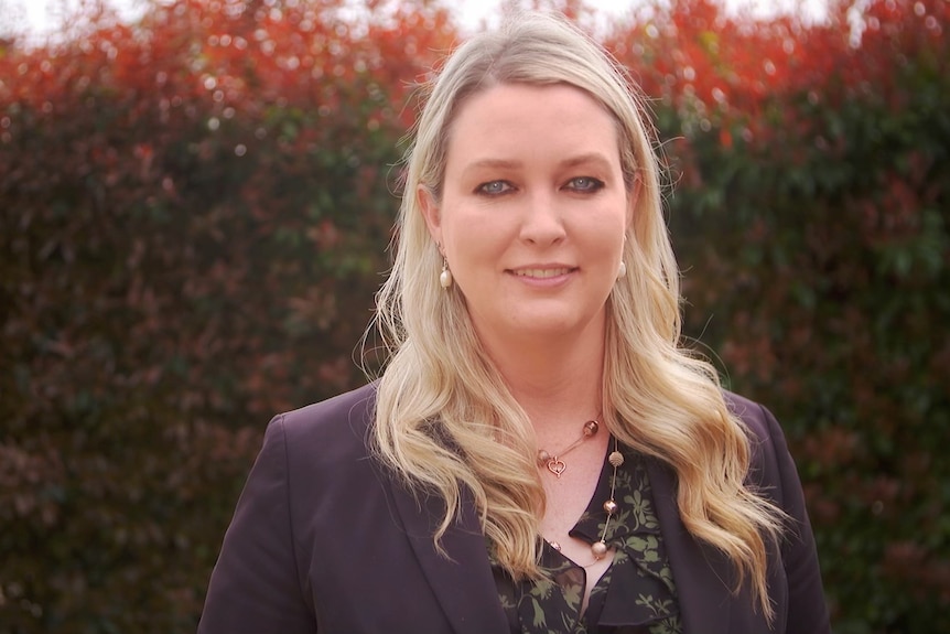 A politician with long blonde hair smiles for the camera outside