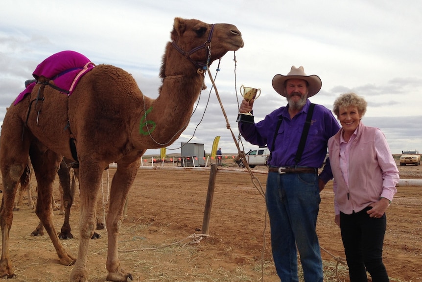 Camel in outback location with male and female standing on red dirt. Male is holding a trophy and a rope attached to the camel. 
