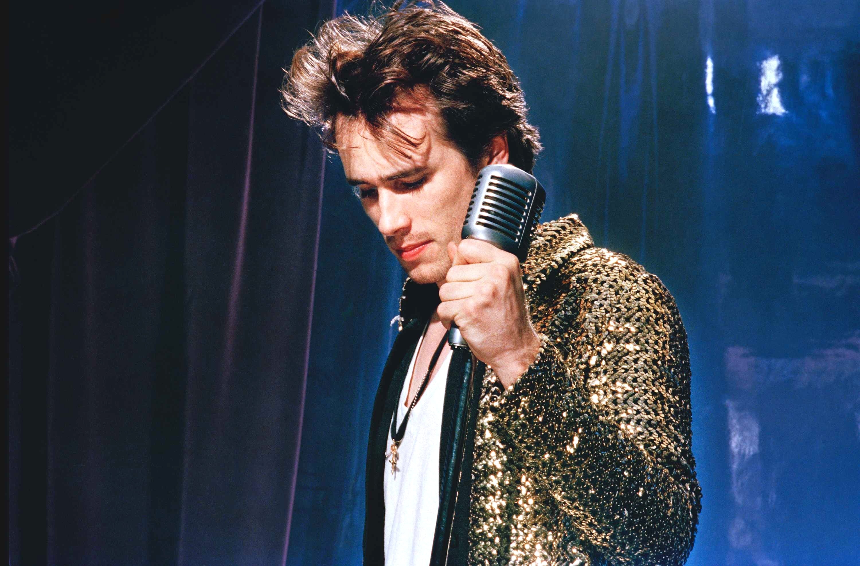 a man in a green plaid jacket starting into the camera, there is a colourful light fixture above him and a mirror to his left