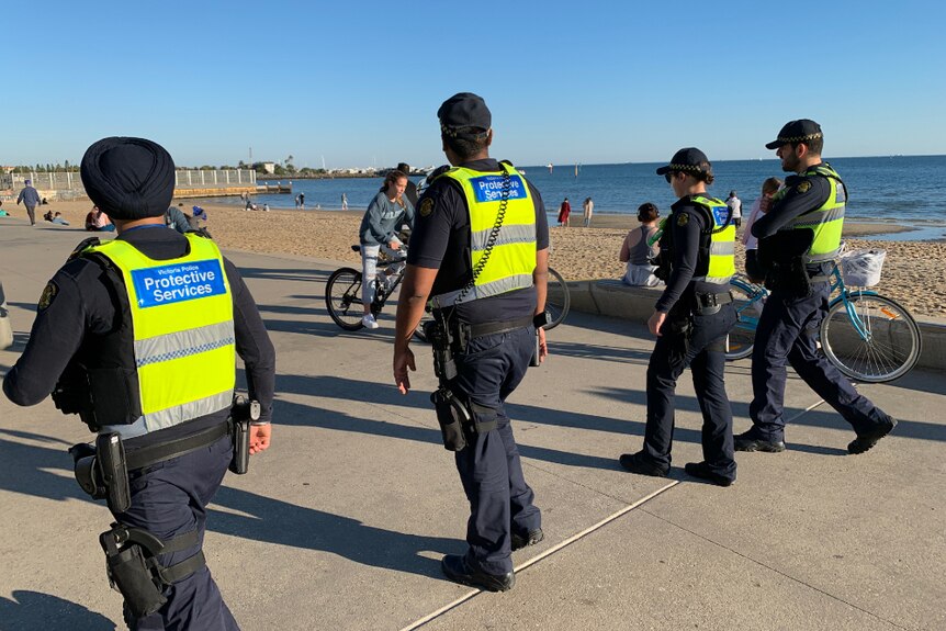 Four protective services officers walk along a path near the beach on a sunny day, with their backs to the camera.