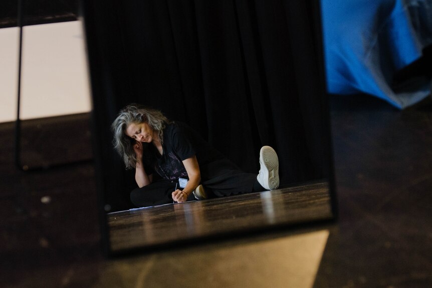 A woman sits with her legs spread out writing on a long sheet of paper on the floor
