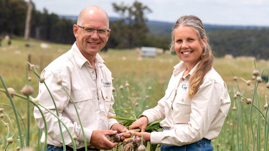 A man and a woman, wearing matching cream-coloured shirts, stand in a paddock and hold garlic.