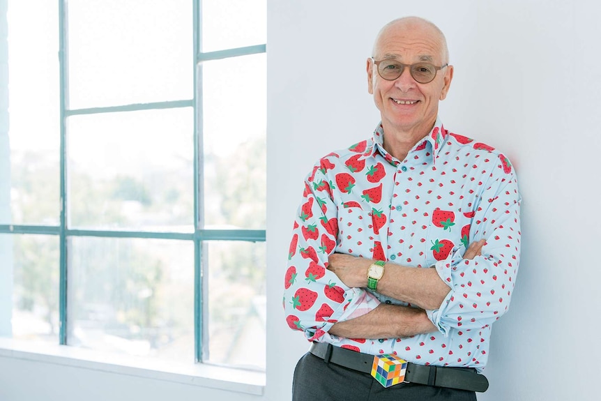 A man wearing a shirt printed with strawberries smiles as he leans against a white wall.