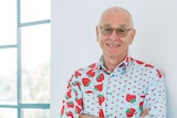 A man wearing a shirt printed with strawberries smiles as he leans against a white wall.
