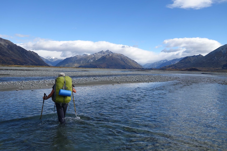 A woman wades through a river against a serene back drop of mountains and blue sky