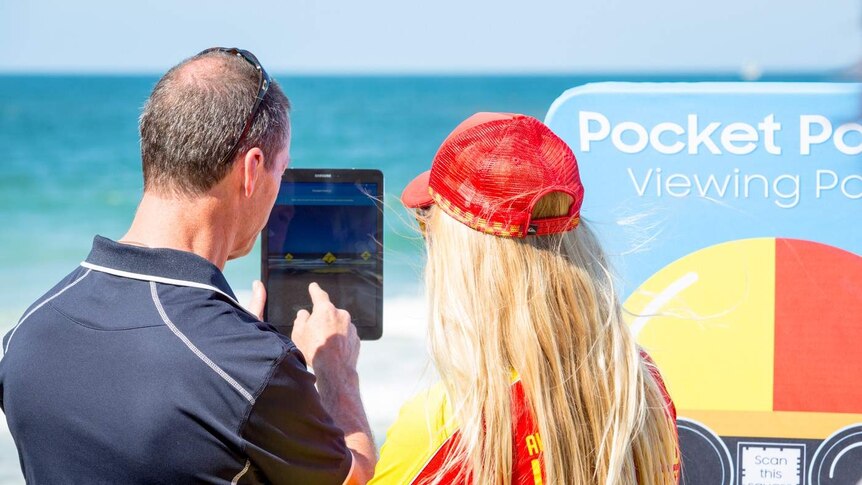 Two people look at a device at the beach and on the screen there's icons showing hazards in the water.