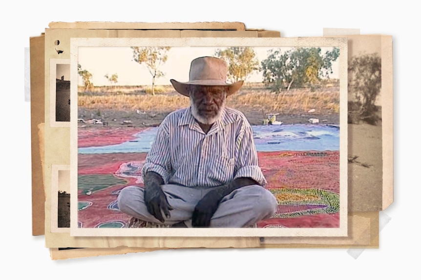 An Aboriginal man with a white beard wearing a cowboy hat sits on a large painted canvas with bush in the background