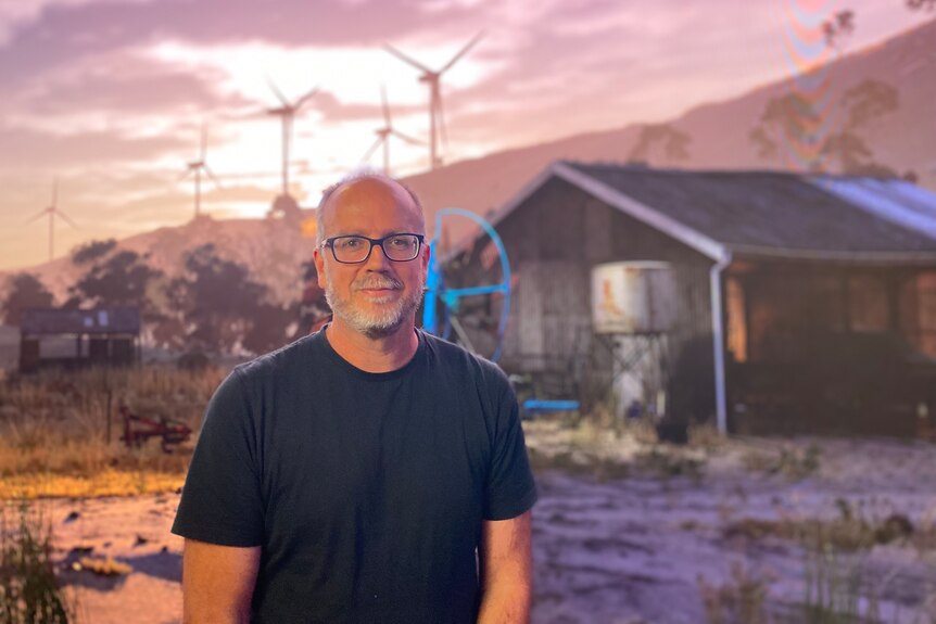 A man wearing black glasses and a black t shirt smiles in front of a digital background of a shed and wind turbines.