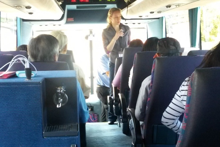 Veronica Macpherson stands at the front of a bus talking into a microphone to passengers.