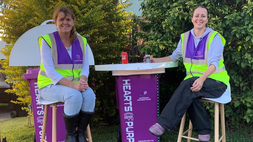 two women sit around a purple wheelie bin with a table top and writing on side of the bin that says hearts of purple