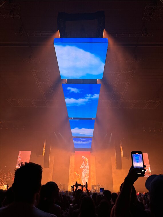 A concert with screens showing blue skies across the ceiling and a man singing on the stage screens