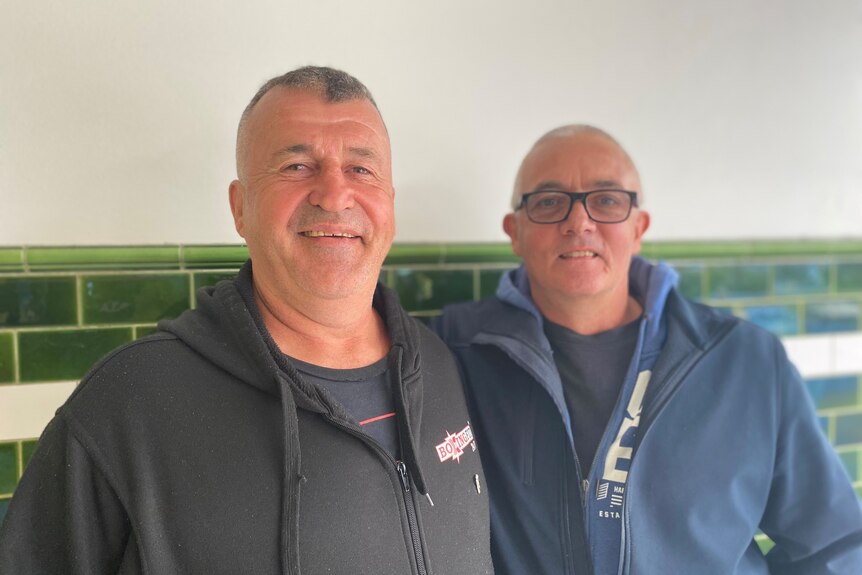 Rod Owen and Glen Fearnett stand in front of a green tiled wall and smile.
