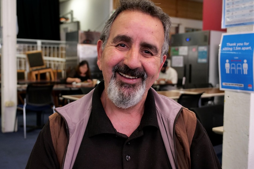 An elderly man with grey facial hair smiling inside a large hall