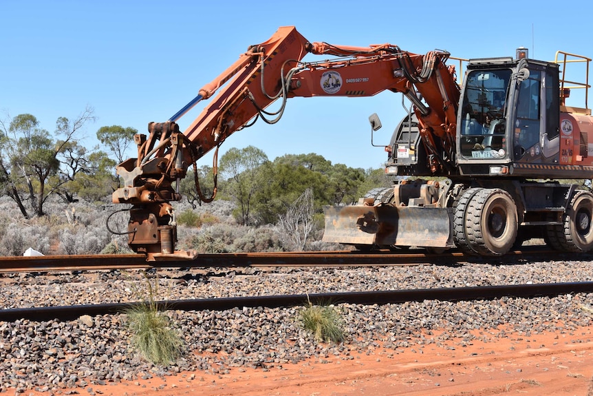 A piece of heavy machinery lifts up a piece of rail.  There is outback red dust surrounding the rail.