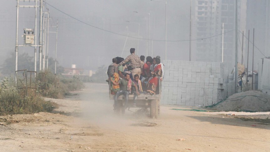 A rickety vehicle overflowing with women, men and children travels on a dirt road, surrounded by air pollution