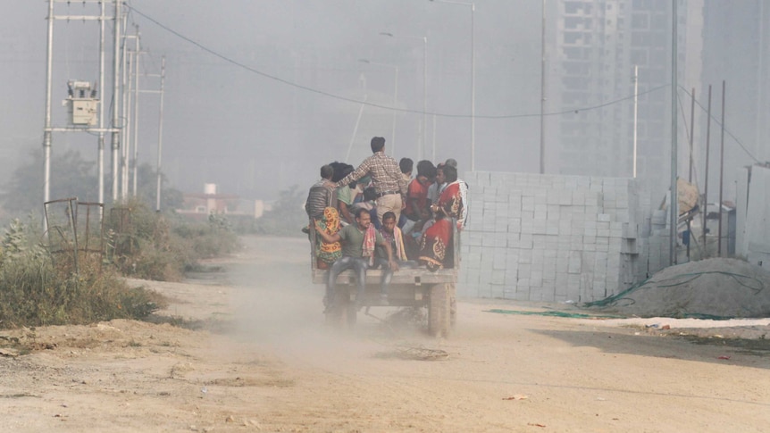 A rickety vehicle overflowing with women, men and children travels on a dirt road, surrounded by air pollution.