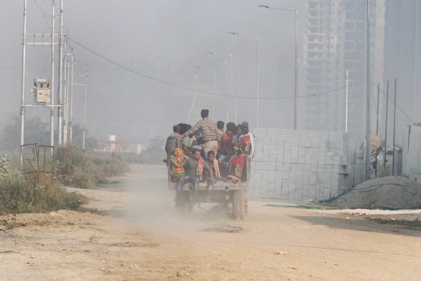 A rickety vehicle overflowing with women, men and children travels on a dirt road, surrounded by air pollution.