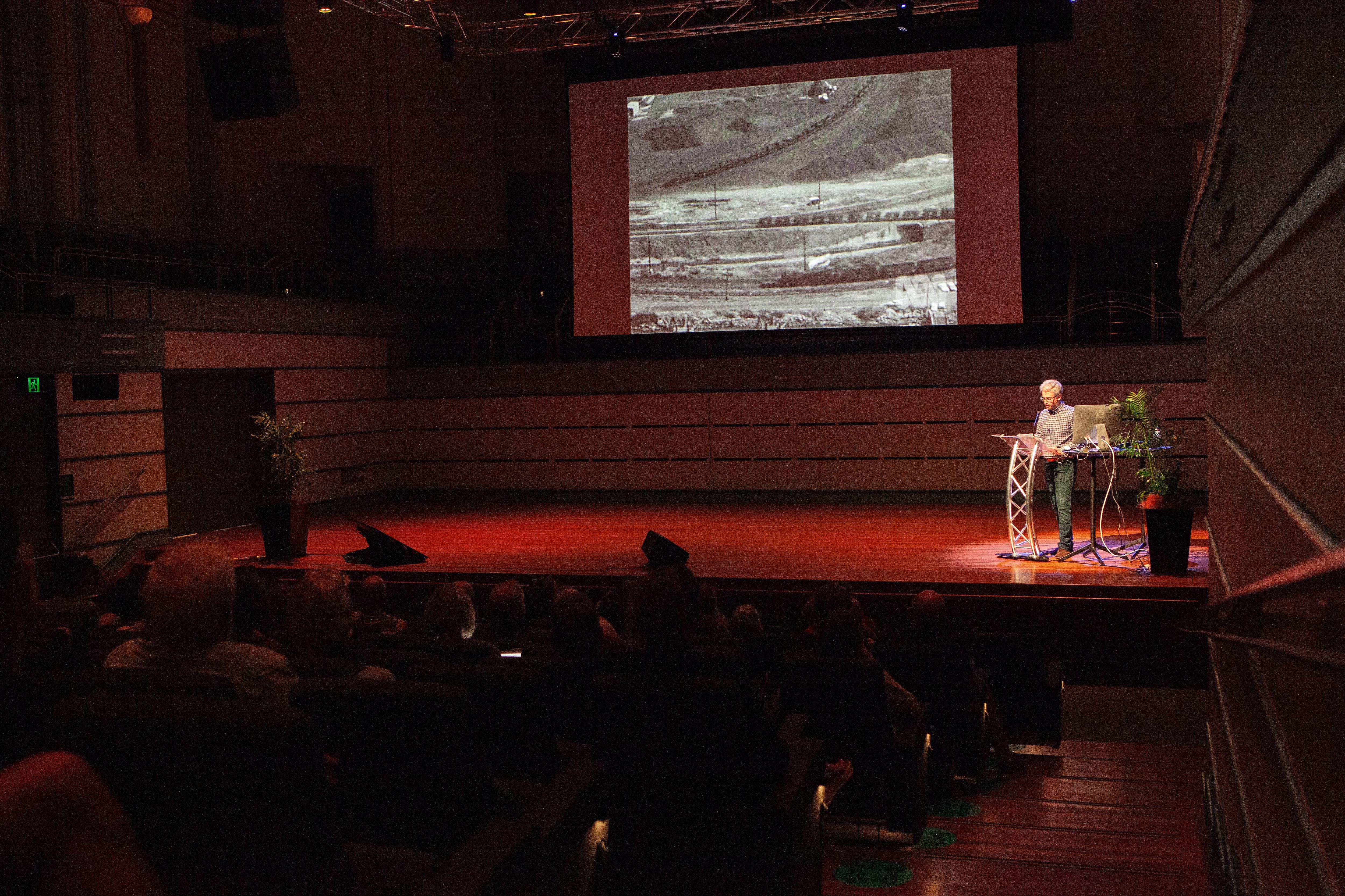 Man on stage at lectern with black and white image of trains on large screen.