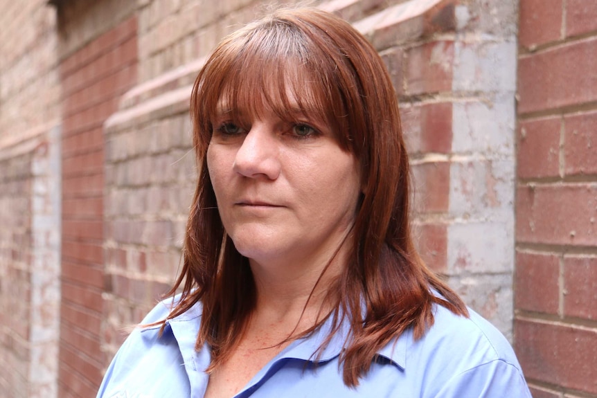 A woman with red hair looks into the distance with a brick wall behind her