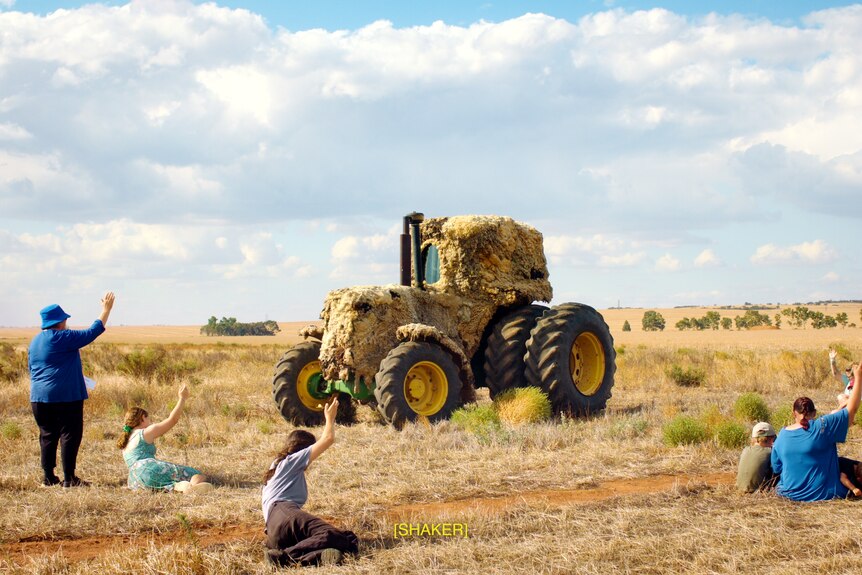 People stand, sit and lie in a dry paddock near a tractor covered in wool.