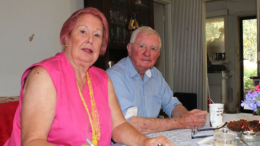 Elderly couple sit side by side at kitchen table and look at camera