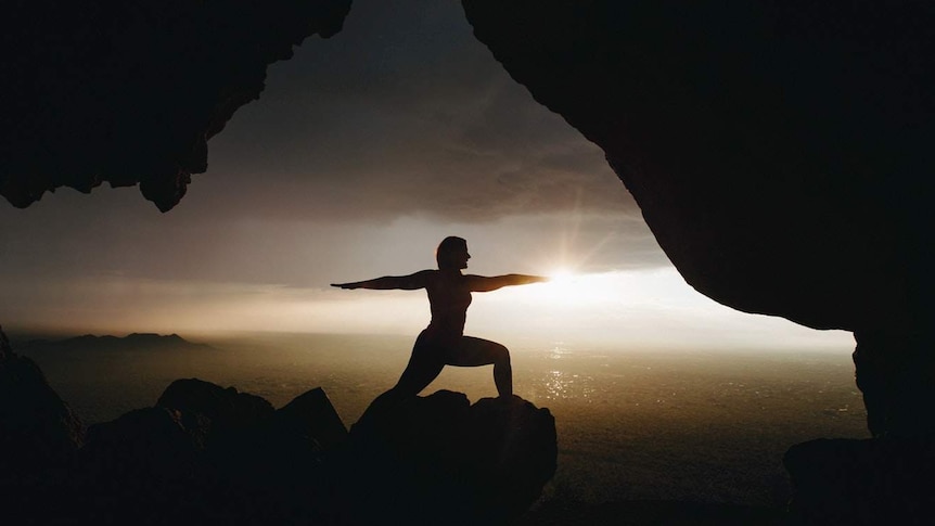 A woman in silhouette does the warrior yoga pose overlooking the ocean, as the sun goes down.