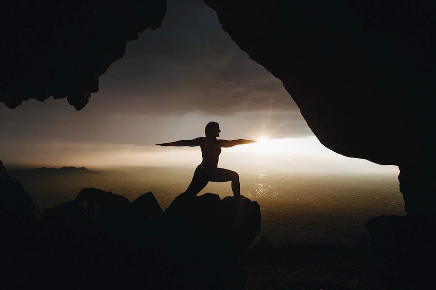 A woman in silhouette does the warrior yoga pose overlooking the ocean, as the sun goes down.
