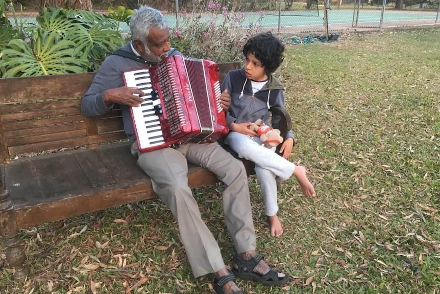 Oscar and his grandfather siting on a park bench playing red accordion. 