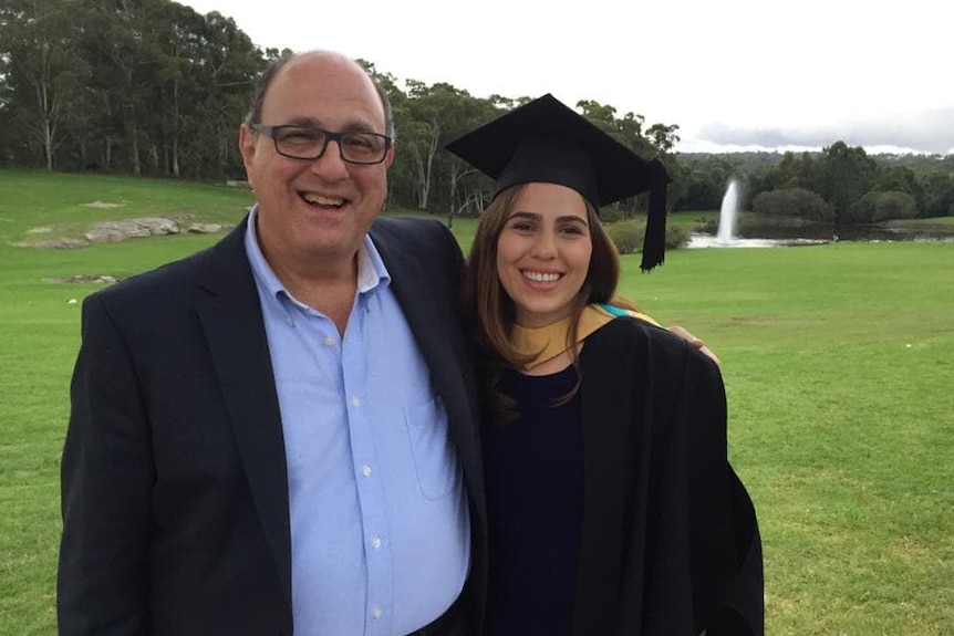A father and daughter stand together on a grassy knoll, smiling at her university graduation.