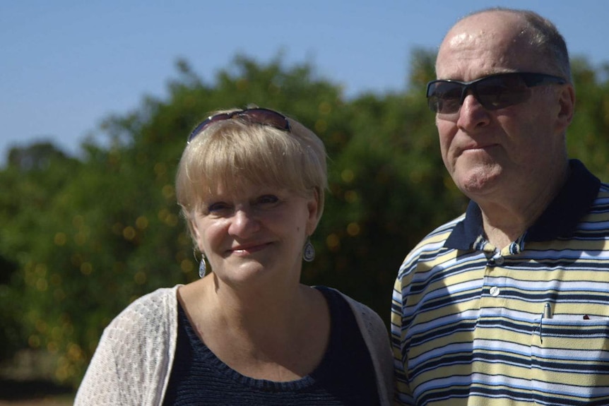 The couple, in their 60s and wearing sunglasses, stand in the sunlight with greenery behind them.