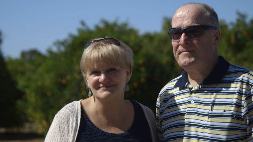 The couple, in their 60s and wearing sunglasses, stand in the sunlight with greenery behind them.